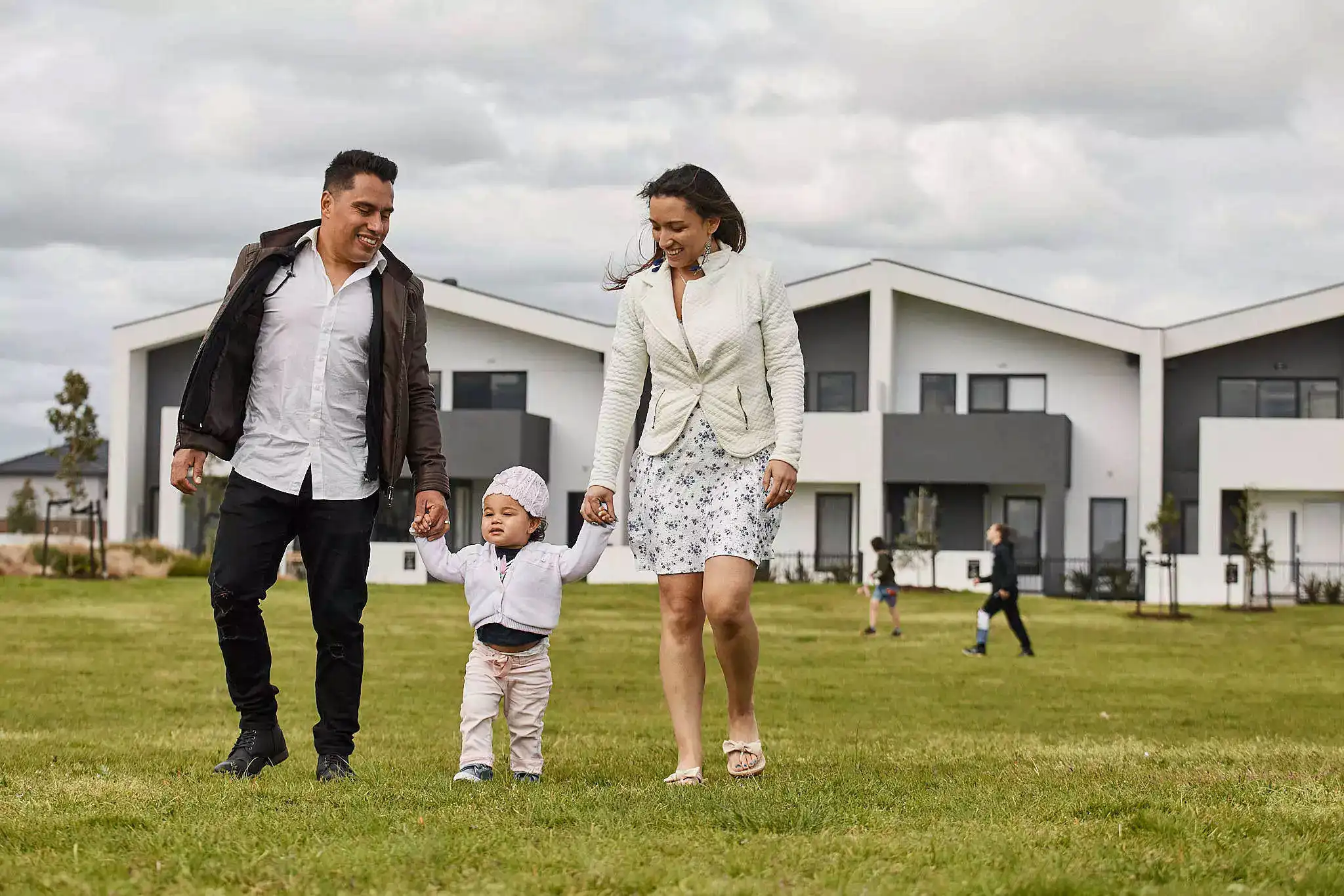 A photograph of a young family walking hand-in-hand across a grassy field with a backdrop of modern, two-storey homes.
