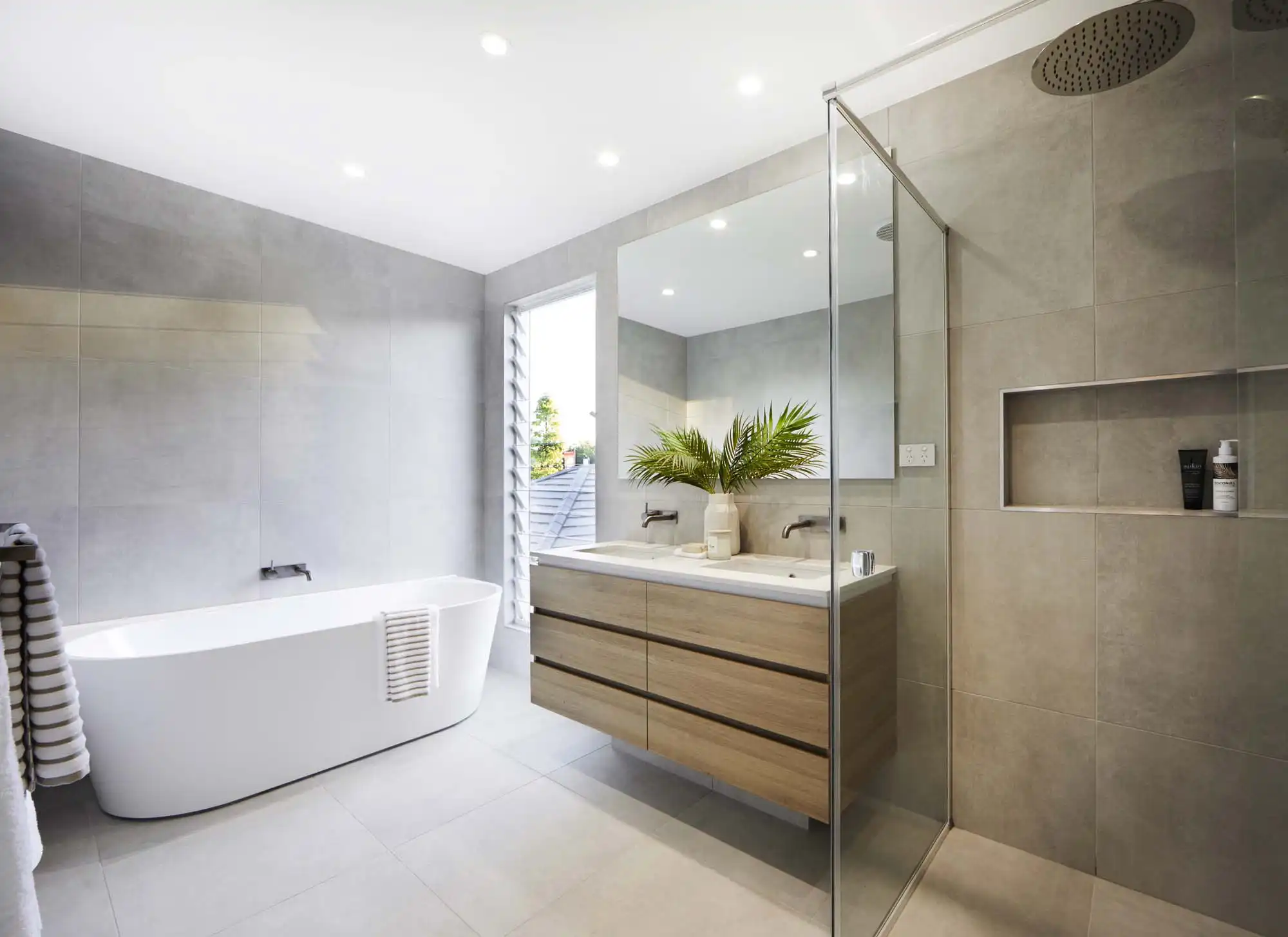 Photo of a bathroom with light grey tiling throughout and a freestanding bath against the wall, as part of the Simonds Queensland home design