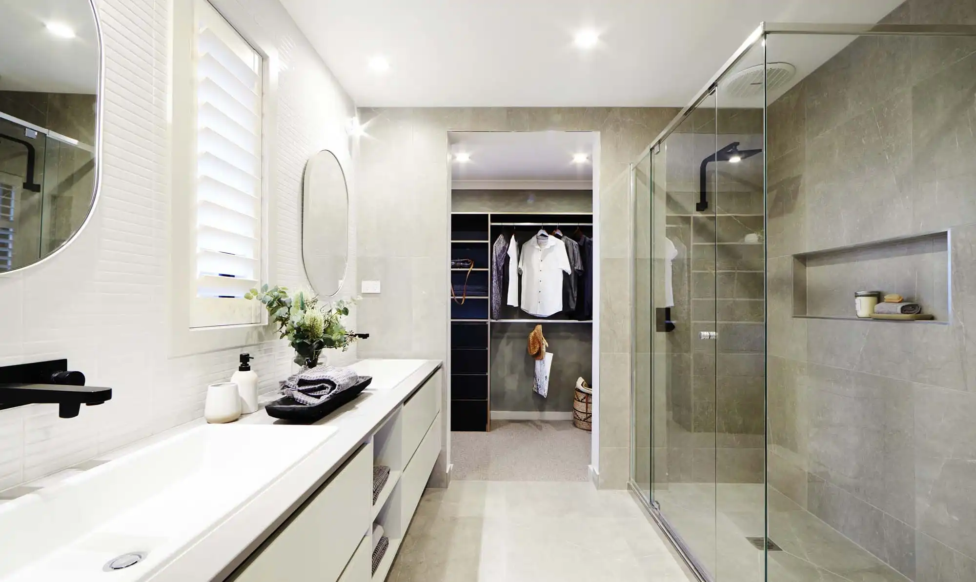 Photo of a bathroom with double vanity featuring white stacked kit kat tiling to the left and shower with glass screen and stone tiles to the right, and through the centre leads into a walk-in wardrobe as part of the Simonds Victoria home design
